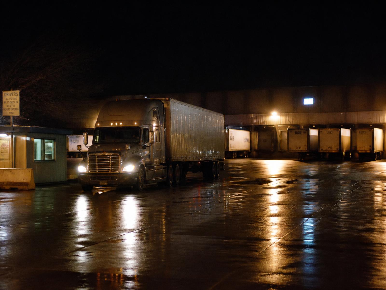 A tractor-trailer leaving an American freight terminal at night, trailers backed into lit loading docks behind it